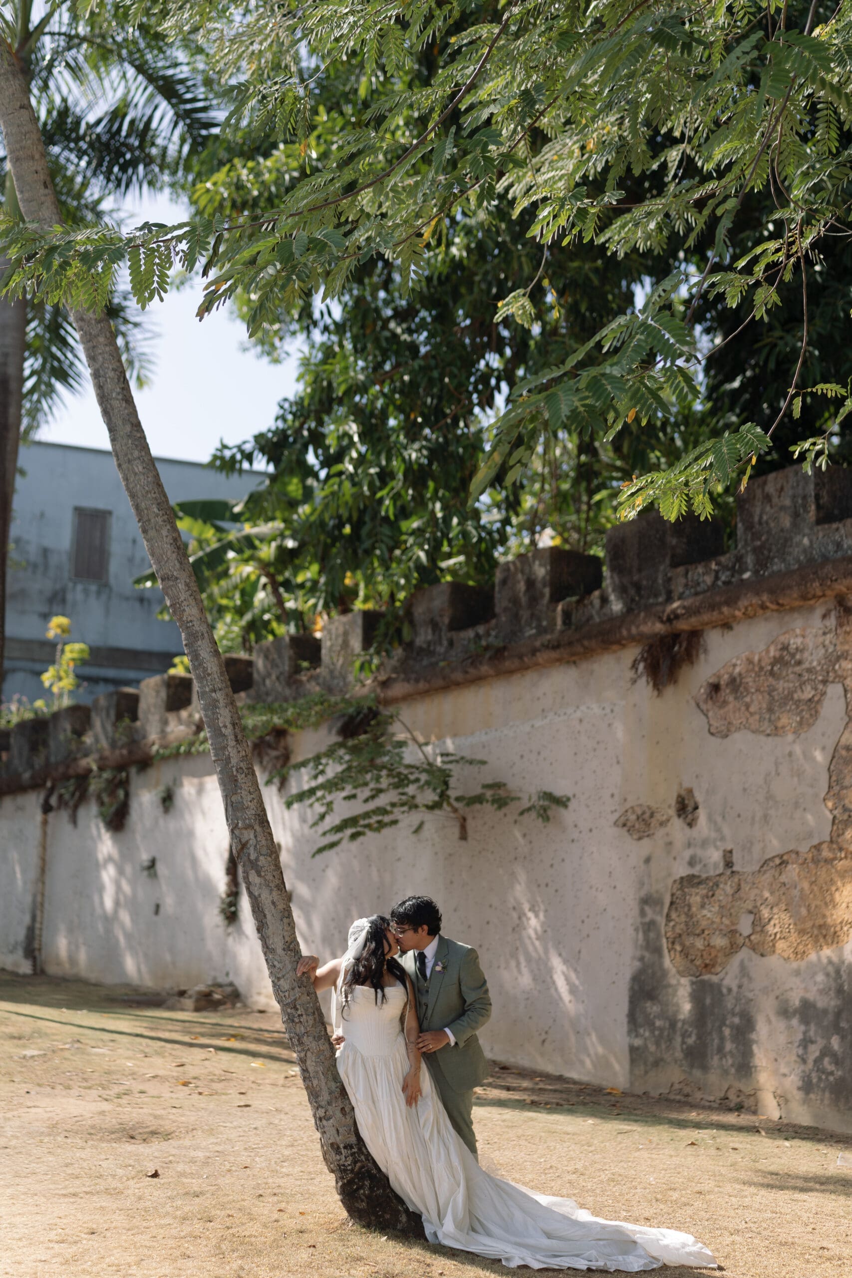 Newlyweds sharing a kiss beneath the trees beside a historic wall in San Juan