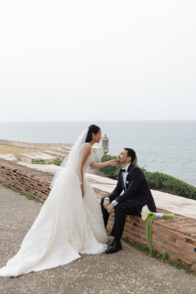 Bride reaches toward the seated groom during a wedding portrait overlooking the ocean at Castillo San Cristóbal.