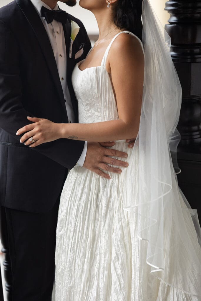 Close-up of the bride and groom embracing, highlighting her textured wedding dress, veil, and wedding rings.
