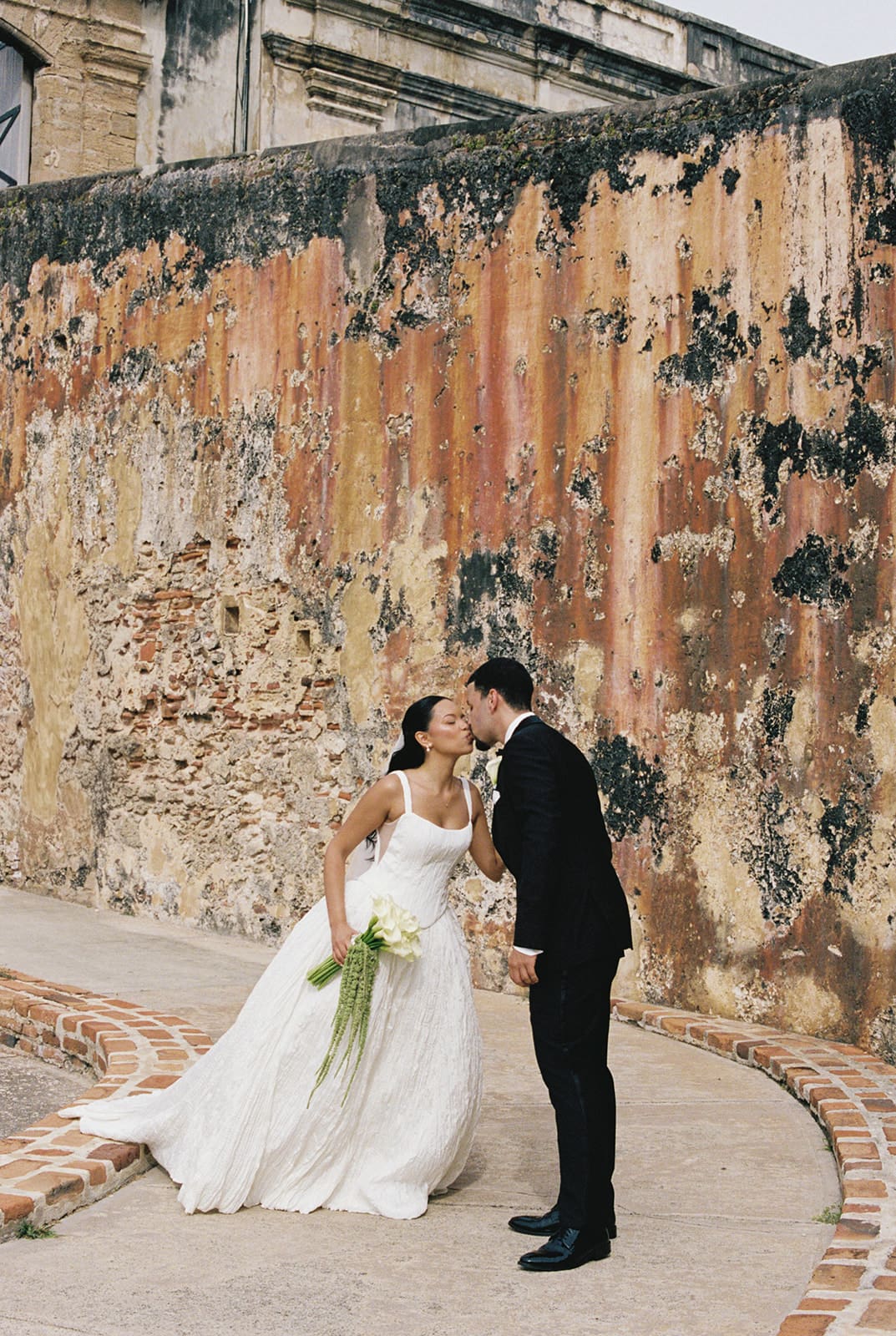 Bride and groom kiss in front of a weathered historic wall at Castillo San Cristóbal in San Juan.