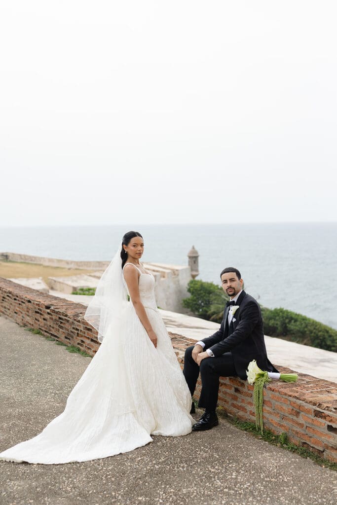 Bride stands beside the seated groom for a wedding portrait overlooking the ocean at Castillo San Cristóbal.