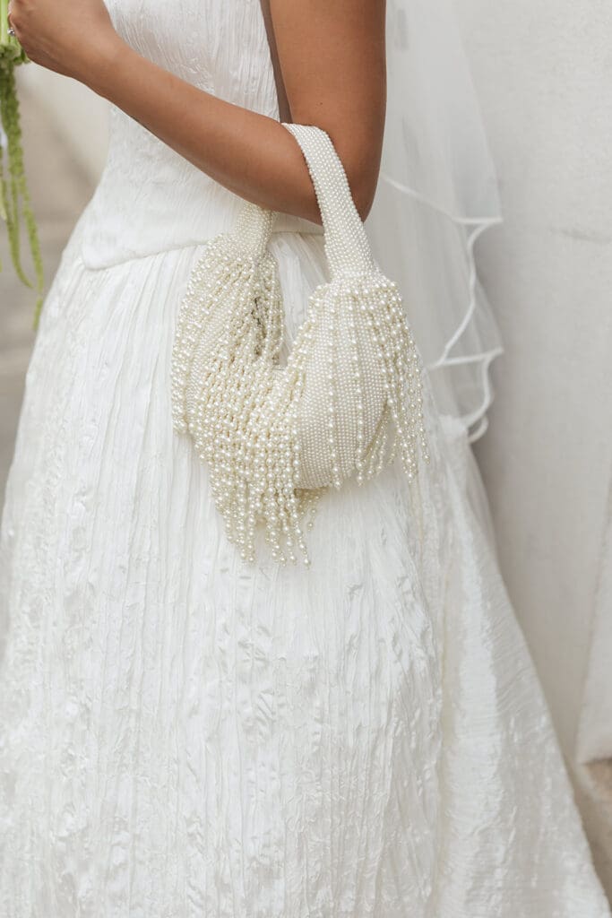 Bride carries a pearl-covered handbag against the textured fabric of her white wedding dress.