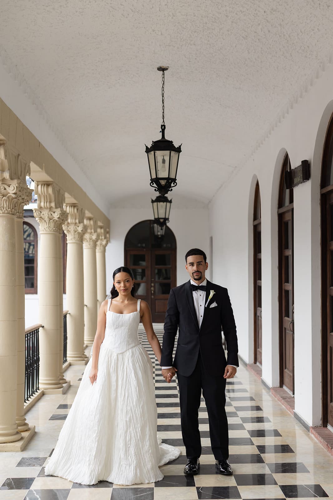 Bride and groom hold hands beneath hanging lanterns in a historic corridor at Casa de España in Puerto Rico.