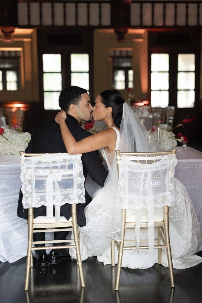 Bride and groom kiss while seated on their decorated “Just Married” chairs at the wedding reception.