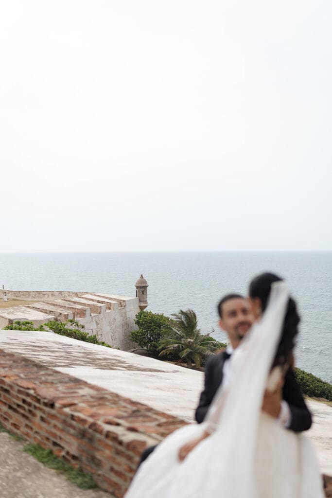 Soft-focus portrait of the bride and groom embracing with the historic fort walls and ocean in the background.