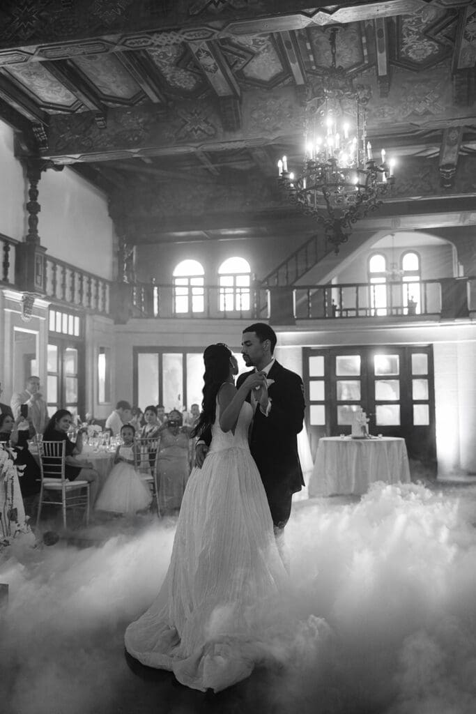 Black-and-white photo of the bride and groom dancing beneath the chandelier with low fog surrounding them.