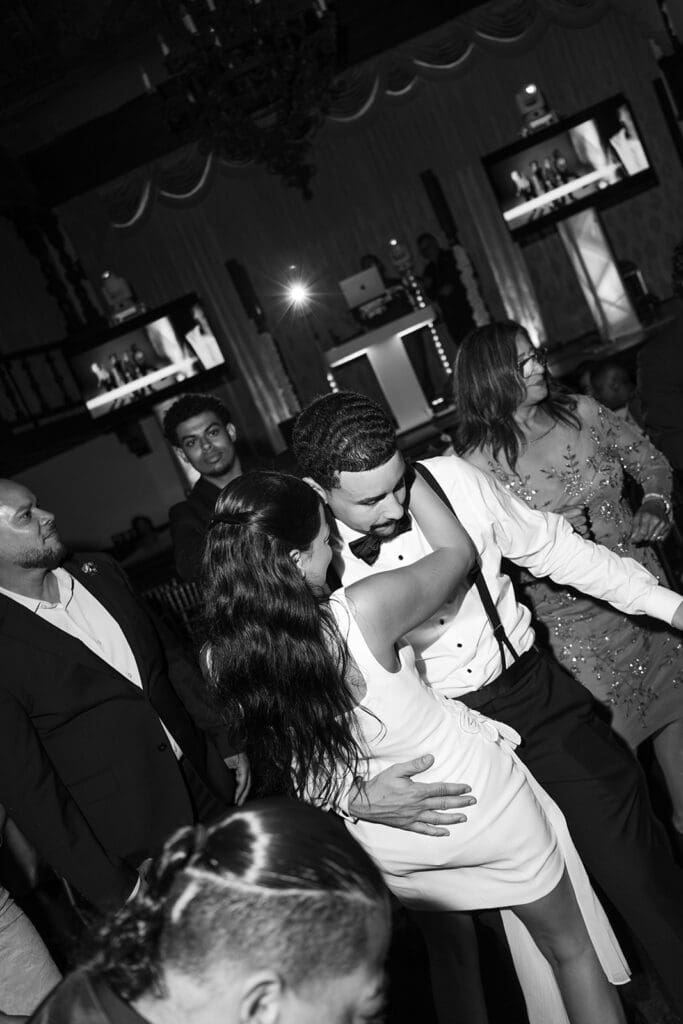 Black-and-white photo of the bride and groom dancing together among guests at their wedding reception.