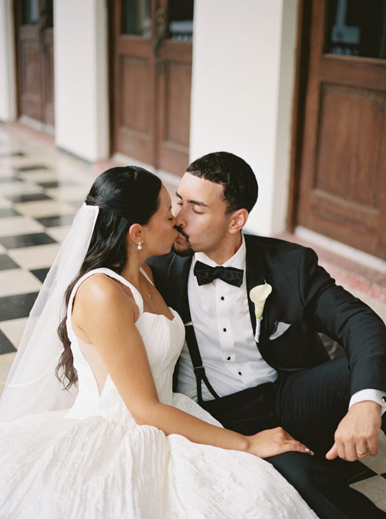 Bride and groom kiss while seated together on the checkerboard floor at Casa de España.