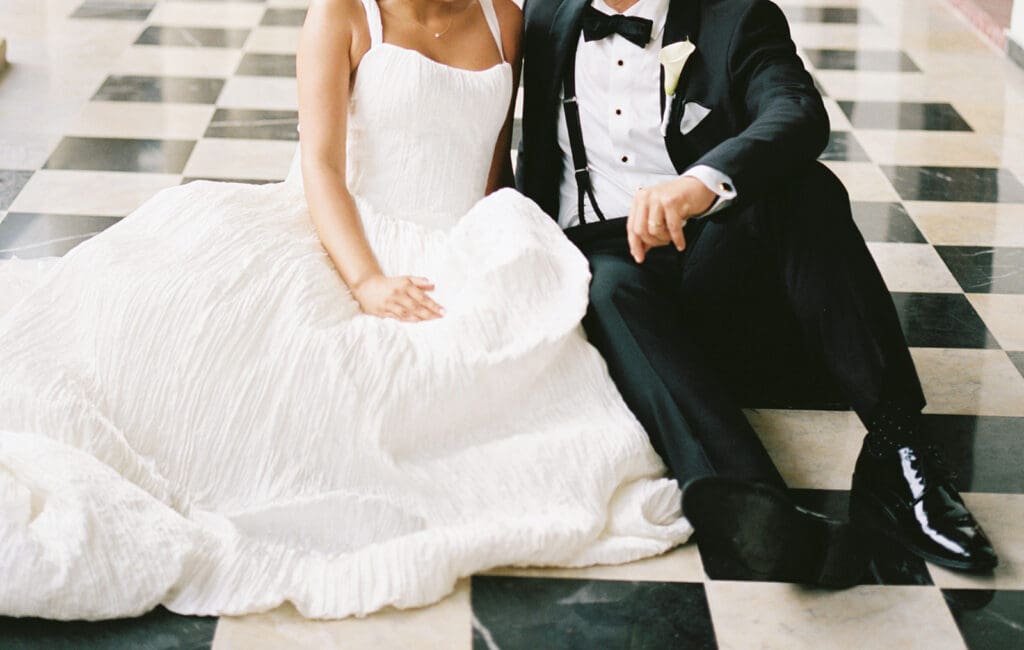 Bride and groom sit together on a black-and-white checkerboard floor, highlighting her textured wedding gown and his tuxedo.