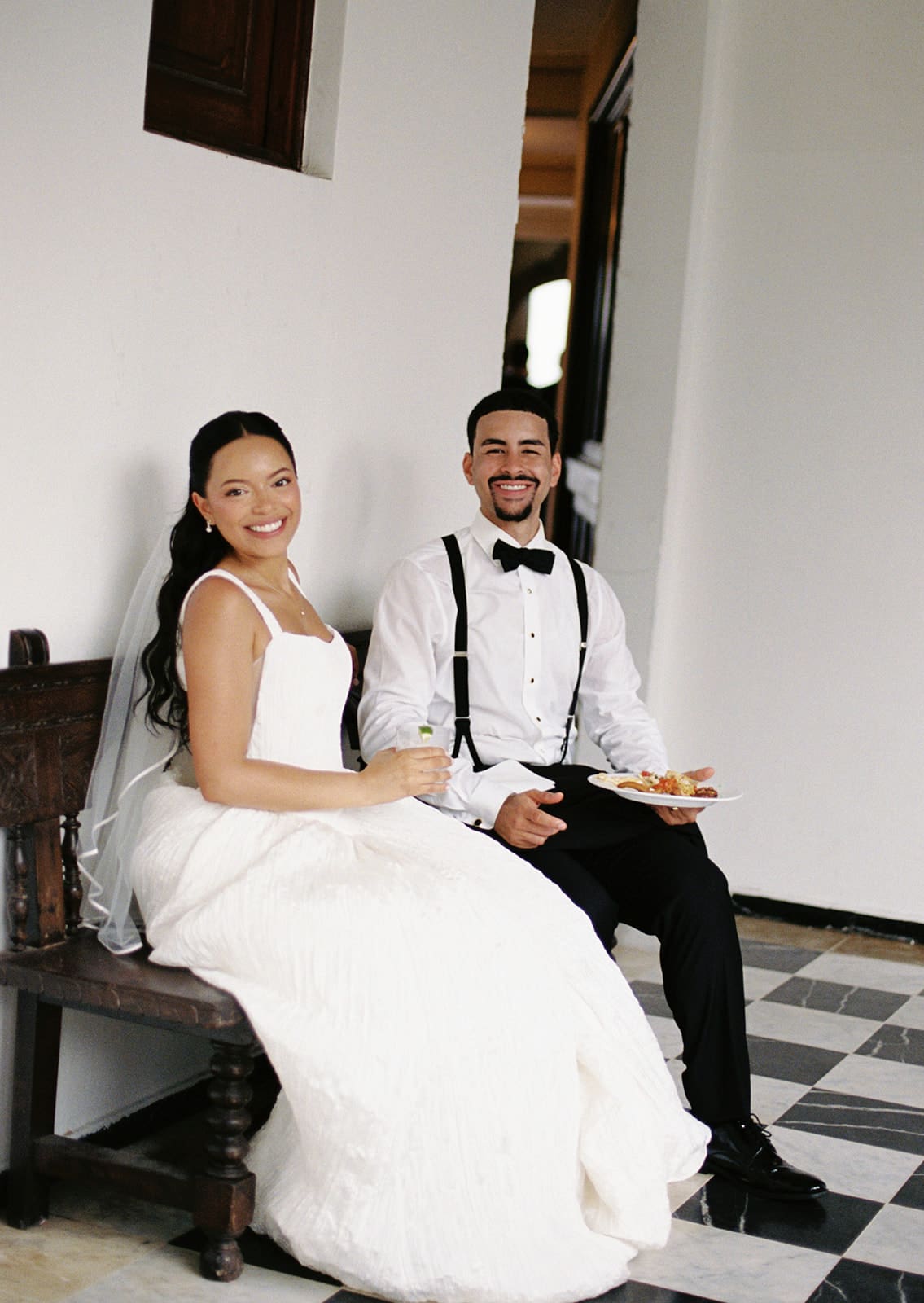 Bride and groom smile together while taking a break on a wooden bench during their wedding celebration.