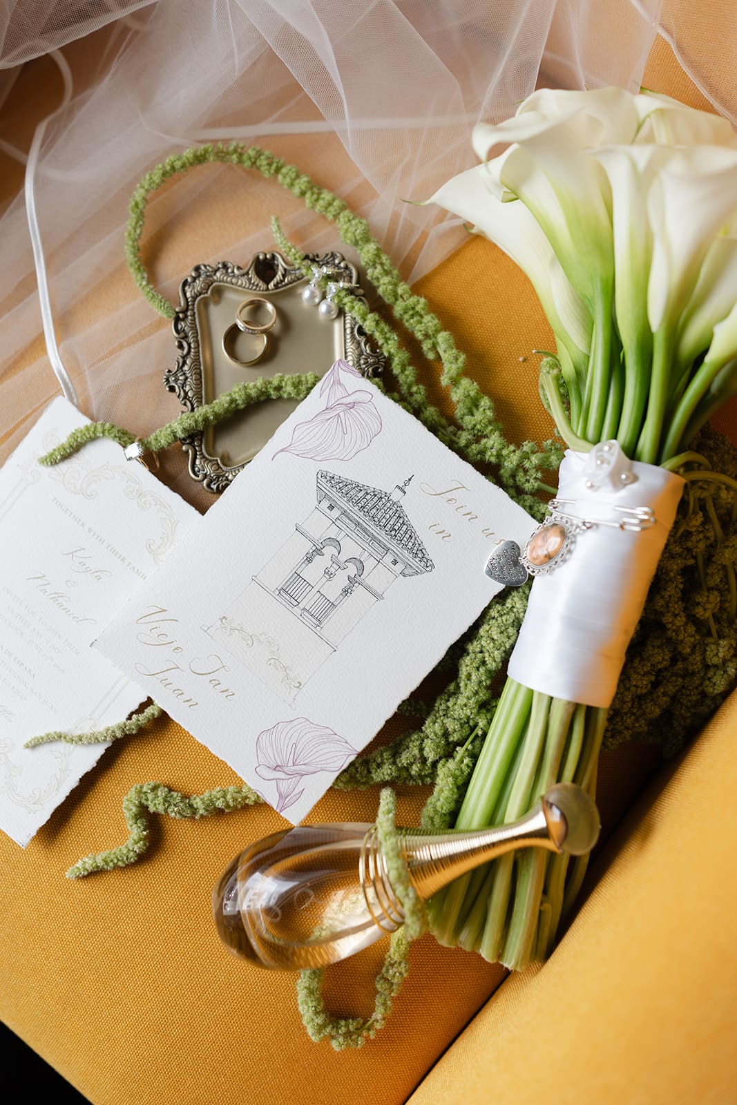 Wedding invitations, rings, pearl earrings and white calla lily bouquet arranged with the bride’s veil.