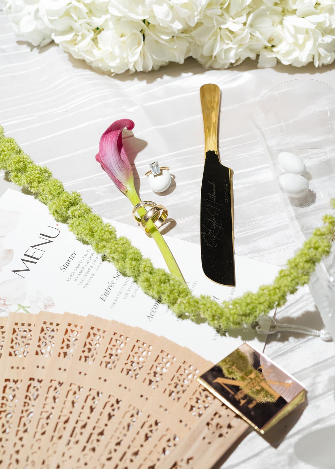 Wedding menu, rings, cake knife, wooden fan, and floral details arranged on the reception table.