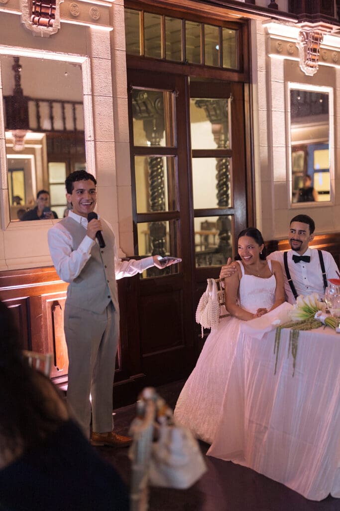Wedding guest giving a speech while the bride and groom laugh at their reception.