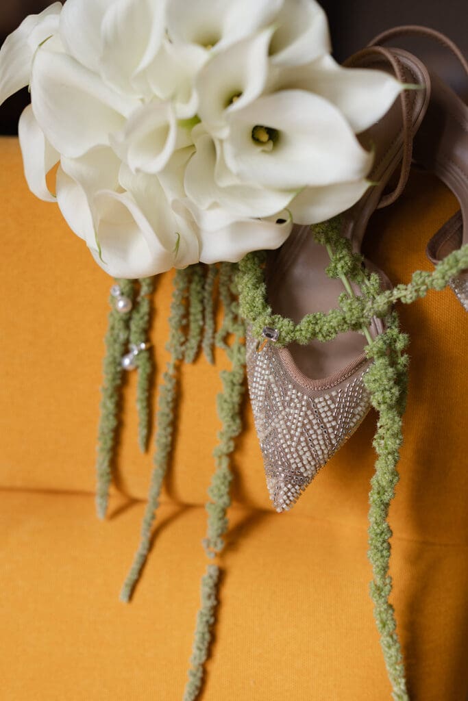 White calla lily bridal bouquet with trailing green flowers beside a beaded wedding shoe.