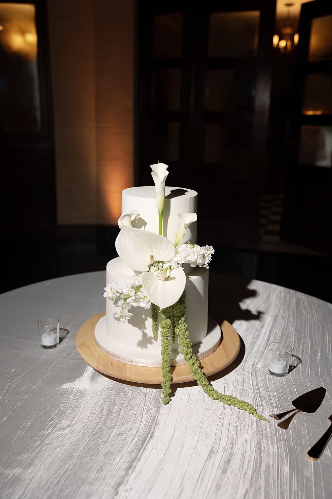 Two-tier white wedding cake decorated with calla lilies, white flowers, and cascading green amaranthus.