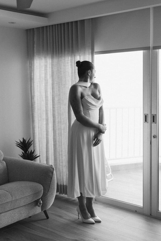 Black and white bridal portrait of a bride in a white midi wedding dress standing beside a window before her Puerto Rico elopement.