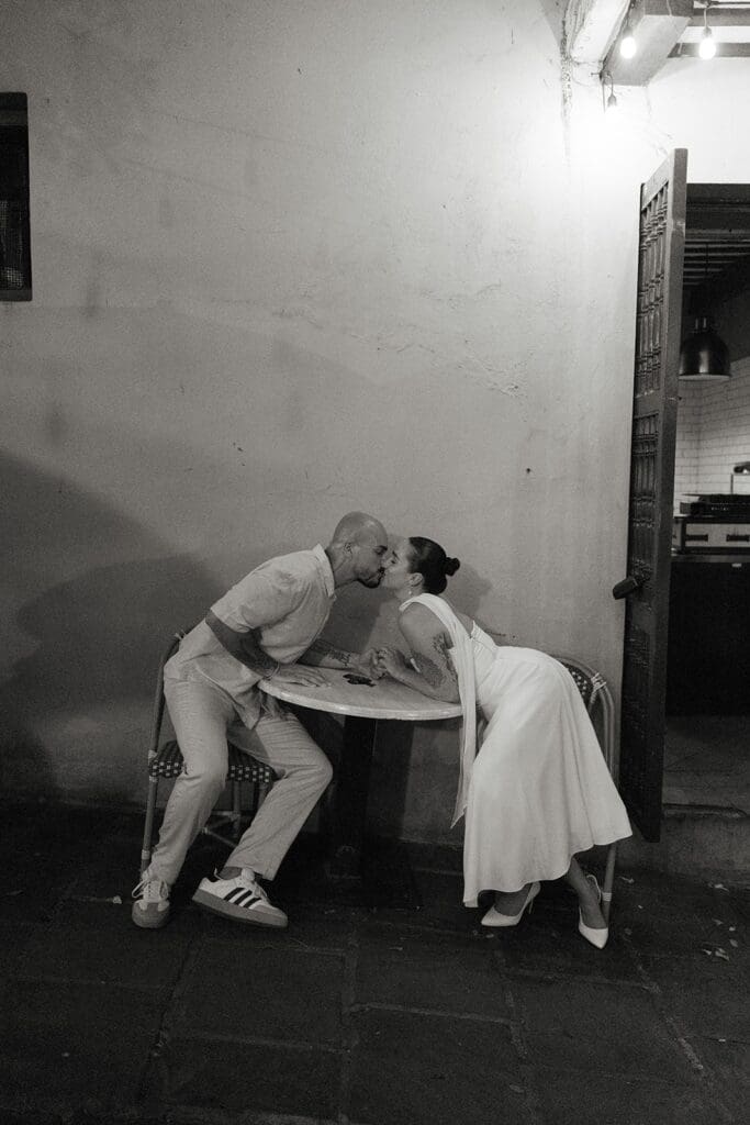 Black and white photograph of a bride and groom leaning across a small café table to kiss during their elopement in Old San Juan, Puerto Rico.