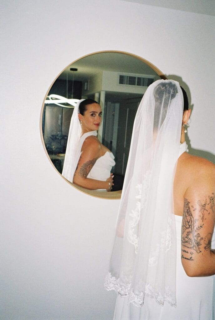 Bride wearing a white wedding dress and lace veil looking at her reflection in a round mirror before her Puerto Rico elopement.