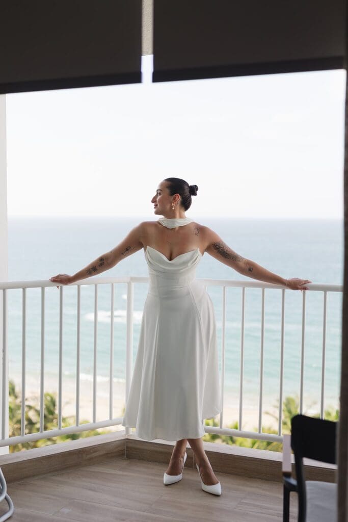 Bride in a white wedding dress and heels standing on a balcony overlooking the ocean before her Puerto Rico elopement.
