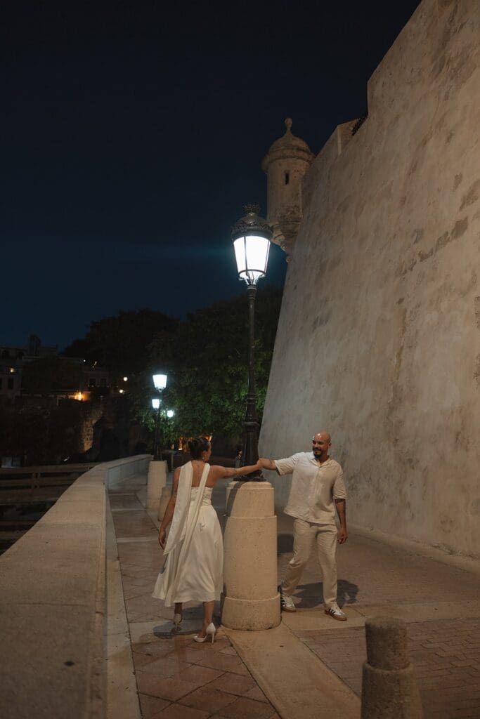 Bride and groom holding hands beside a glowing streetlamp along the waterfront fortress walkway in Old San Juan at night.