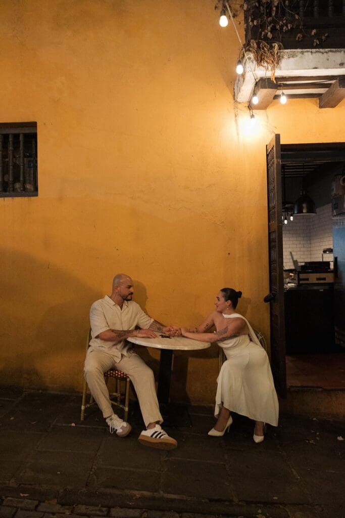 Bride and groom sitting across from each other at a small outdoor café table beside a yellow wall in Old San Juan during their Puerto Rico elopement.