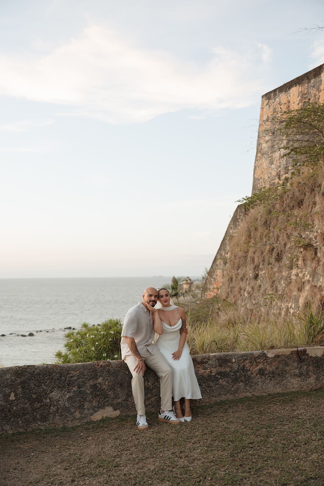 Bride and groom sitting together on a historic stone wall overlooking the ocean during their Puerto Rico elopement in Old San Juan.