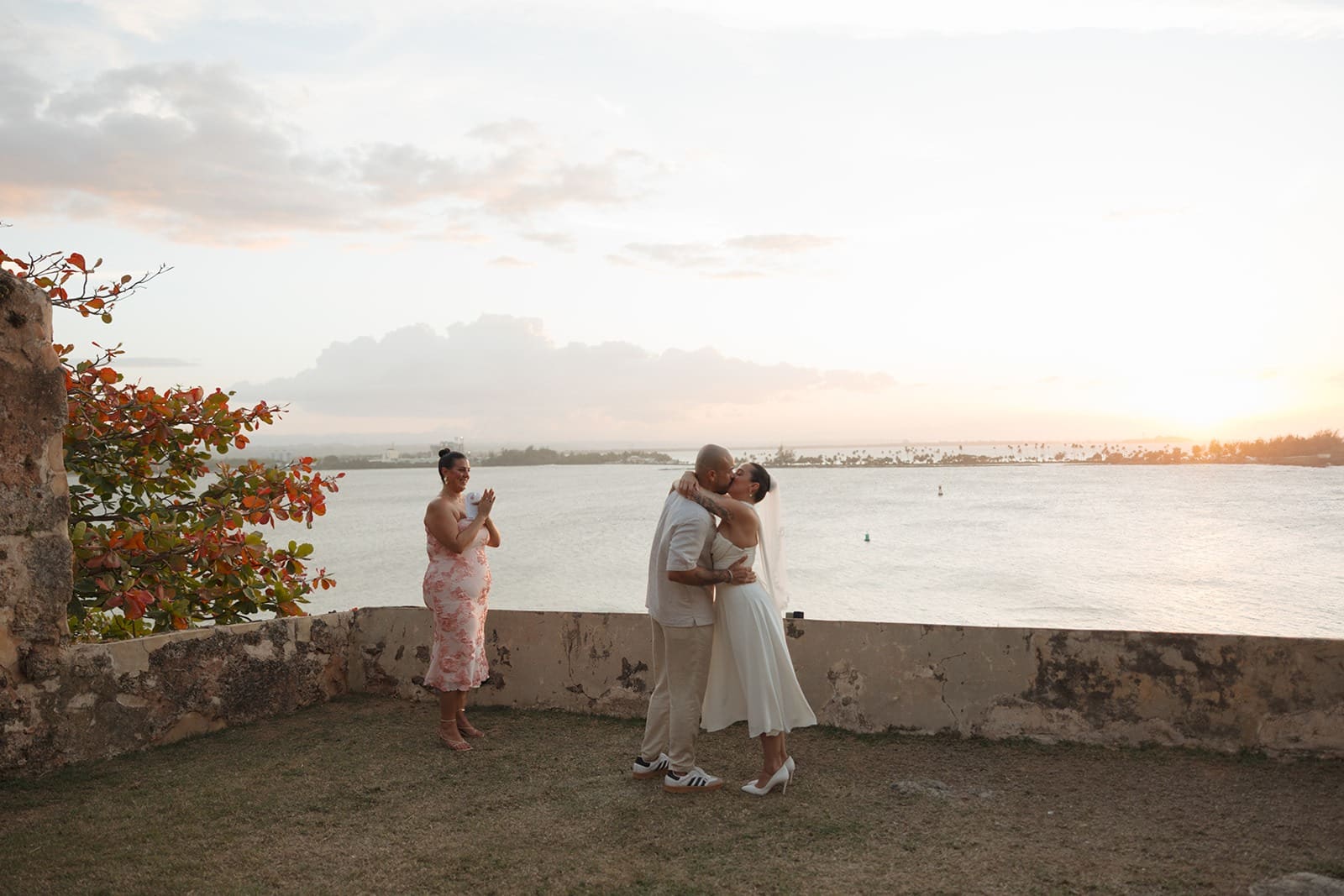 Couple sharing a kiss during their Puerto Rico elopement ceremony at sunset, with the ocean behind them and a guest applauding nearby.
